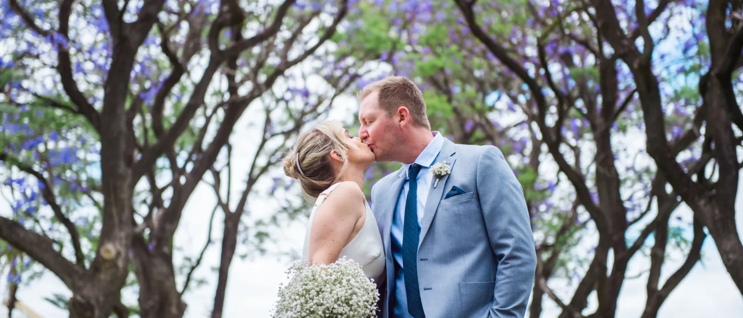 Photo of lizzie and Craig kissing under the Jacaranda trees in full bloom in Hyde Park Perth